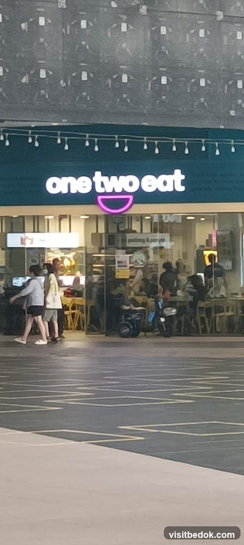 The exterior of a restaurant with the name "one two eat" displayed in large white letters above the entrance. A pink graphic resembling a bowl or mouth is below the text. The restaurant has large glass windows and doors, and people can be seen inside dining. There are also some scooters parked outside the entrance.
