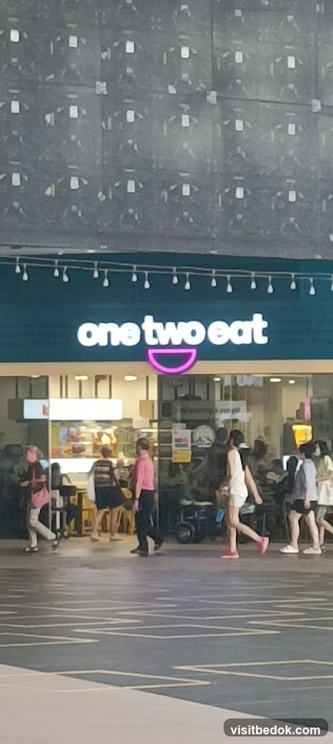 The exterior of a restaurant with a teal facade and a neon sign that reads "one two eat" with a pink bowl graphic. People are walking past the entrance, and the interior of the restaurant is visible, showing food displays and seating areas.