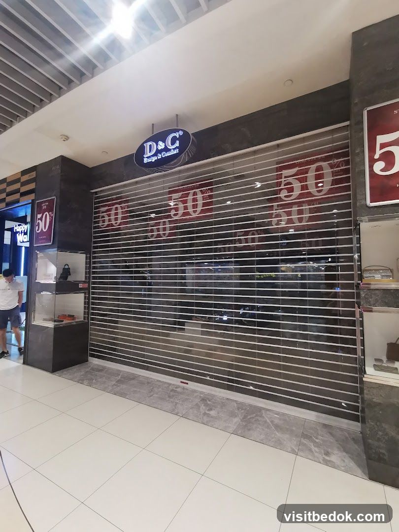 The storefront of a closed shop with a transparent roll-down gate. A sign above the gate reads "D&C Design & Comfort". Red signs with "50%" are visible on the gate, indicating a sale. To the left, a display case shows some items, and a person is walking by. To the right, another display case is partially visible.