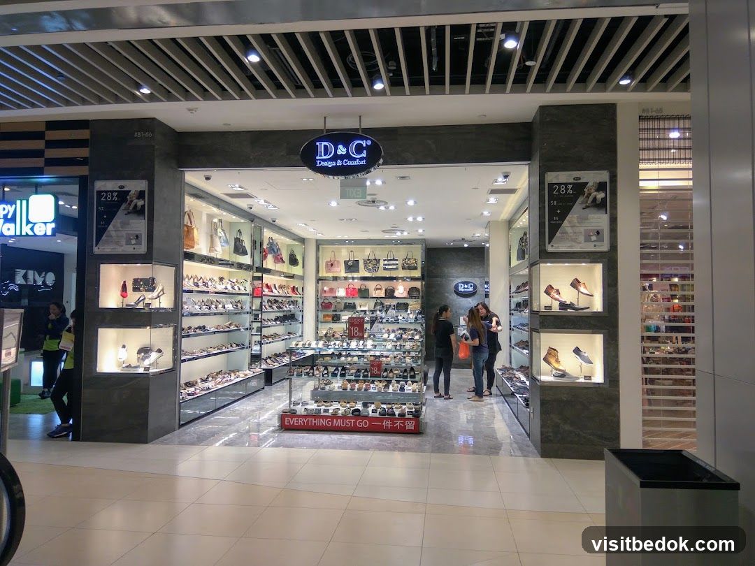 A well-lit shoe store with a wide selection of women's handbags and shoes displayed in glass cases and on shelves. The store features a prominent D&C Design & Comfort sign above the entrance. Promotional posters are visible on the exterior walls.