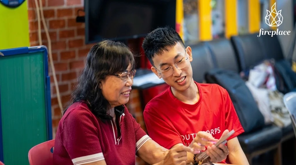 A Youth Corps volunteer in a red shirt sitting with an older woman as they look at a smartphone together