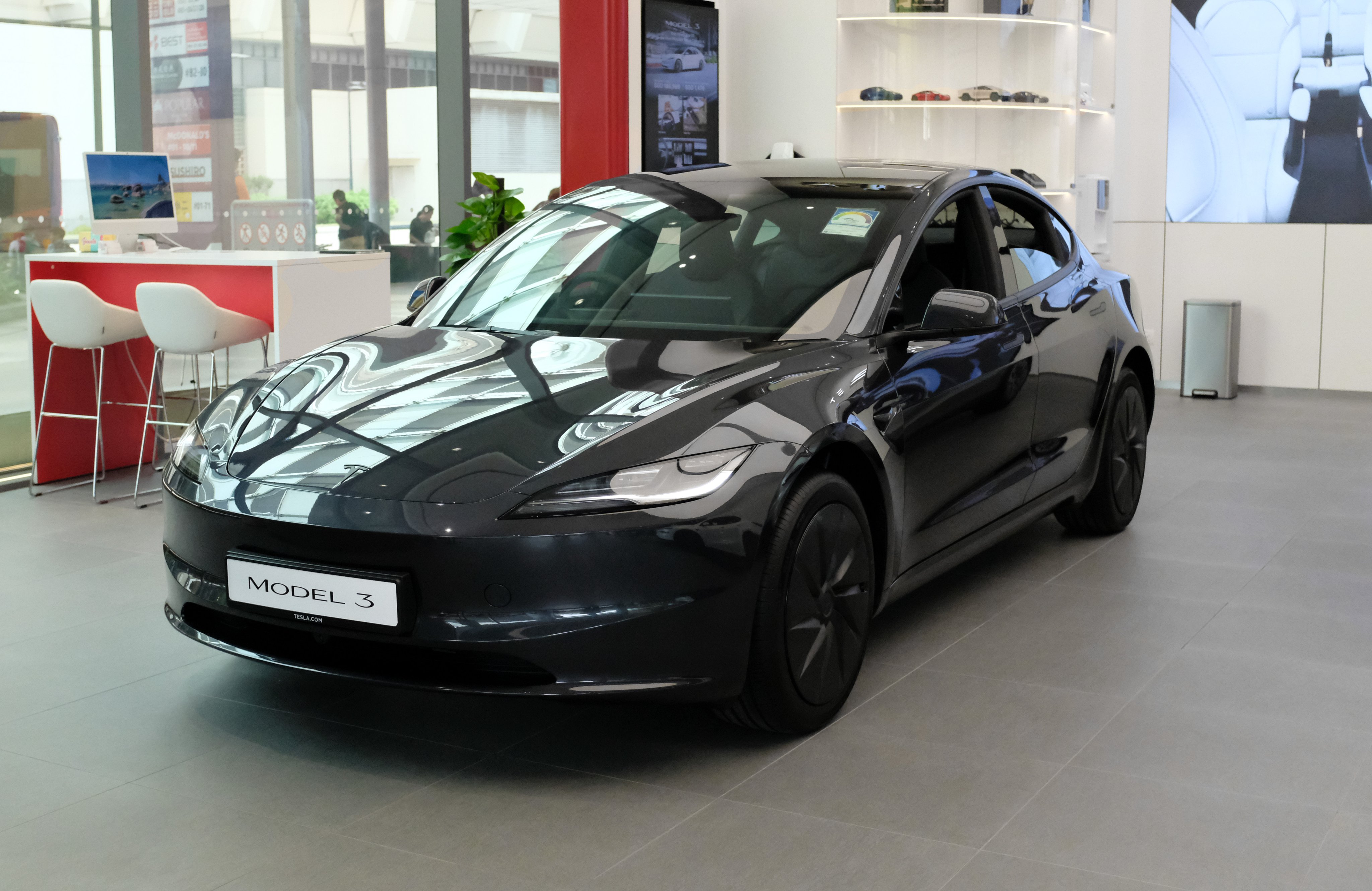 Black Tesla Model 3 inside the Bedok Mall Experience Centre, with the mall atrium visible through the glass